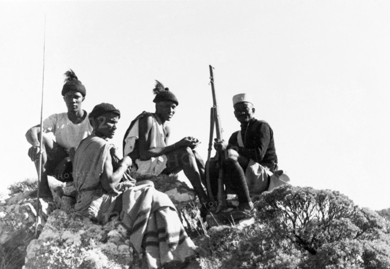 Kikuyu Home Guards on patrol. Four Kikuyu Home Guards on patrol sit atop a rocky outcrop. One wears a uniform and carries a rifle, another holds a spear. Kenya, circa 1952. Kenya, Eastern Africa, Africa.