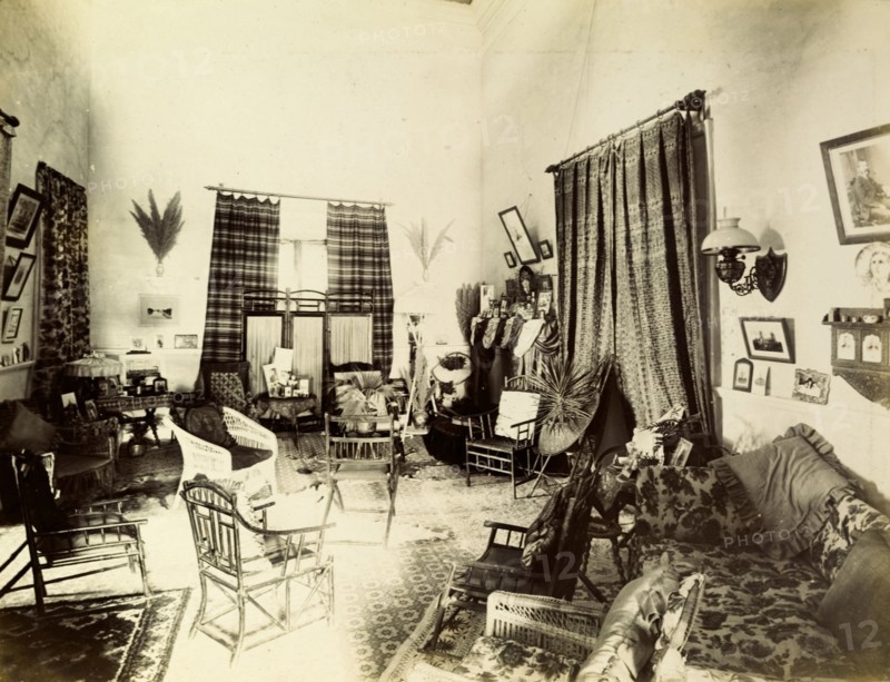 Mrs Morton's drawing room. Interior shot of a traditionally Victorian drawing room belonging to 'Mrs Morton'. Characterised by a high ceiling and floor-length windows, the room is busily decorated with furniture, framed photographs and heavily patterned fabrics. India, circa 1895. India, Southern Asia, Asia.
