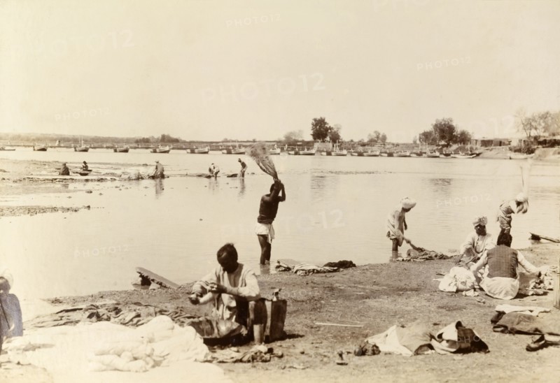 Washing clothes in the river. Indian men wash items of clothing by thrashing them onto special stones at the edge of a wide river. India, circa 1895. India, Southern Asia, Asia.