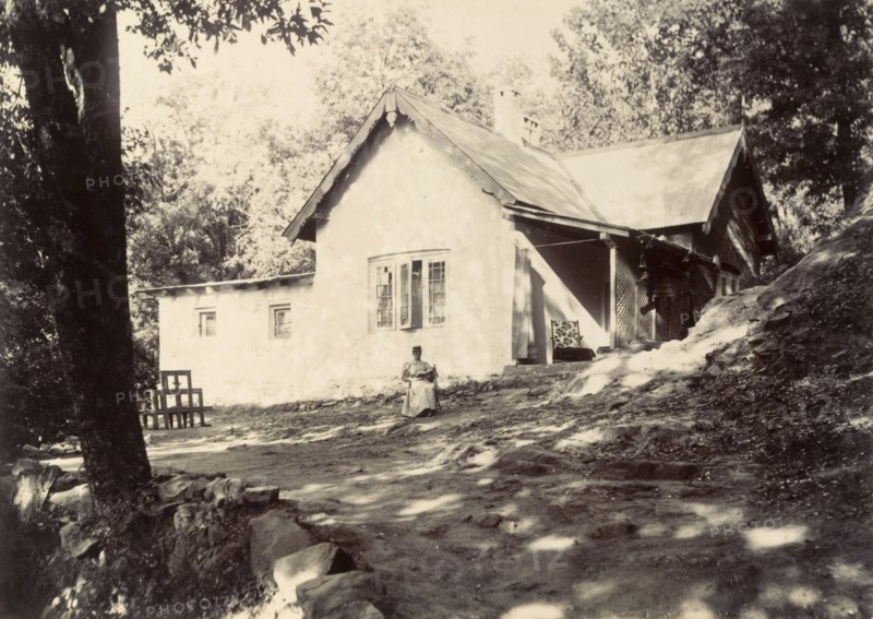 Mrs Page reads in the garden. A distant figure identified as 'Mrs Page' sits reading her book in front of a bungalow. The original caption suggests she is 'watching tongas' (two-wheeled carriages drawn by horses). India, circa 1890. India, Southern Asia, Asia.