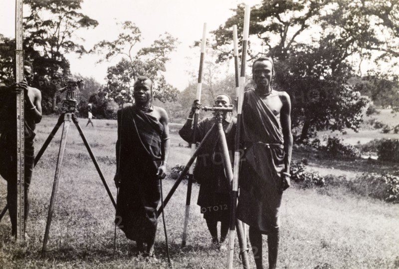 Maasai men help a surveying team. Four Maasai men in traditional dress help a surveying team by holding up distance markers for a hidden photographer. Probably Tanganyika Territory (Tanzania), circa 1930. Tanzania, Eastern Africa, Africa.