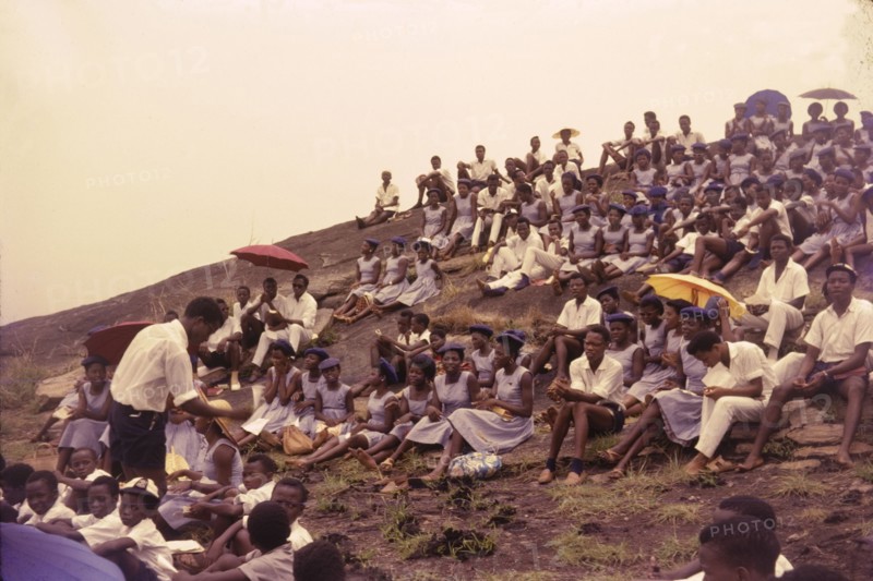 Student union picnic, Nigeria. Uniformed students from a Church of Nigeria college sit on a hillside to enjoy a student union picnic. The men are dressed in white whilst their female counterparts wear blue dresses and berets. Nigeria, circa 1963. Nigeria, Western Africa, Africa.