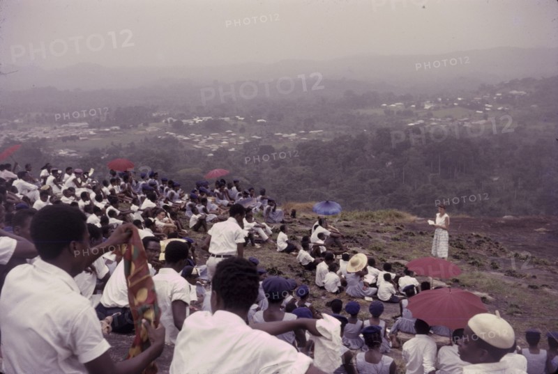 Student union picnic, Nigeria. Uniformed students from a Church of Nigeria college sit on a hillside to enjoy a student union picnic. The men are dressed in white whilst their female counterparts wear blue dresses and berets. Nigeria, circa 1963. Nigeria, Western Africa, Africa.