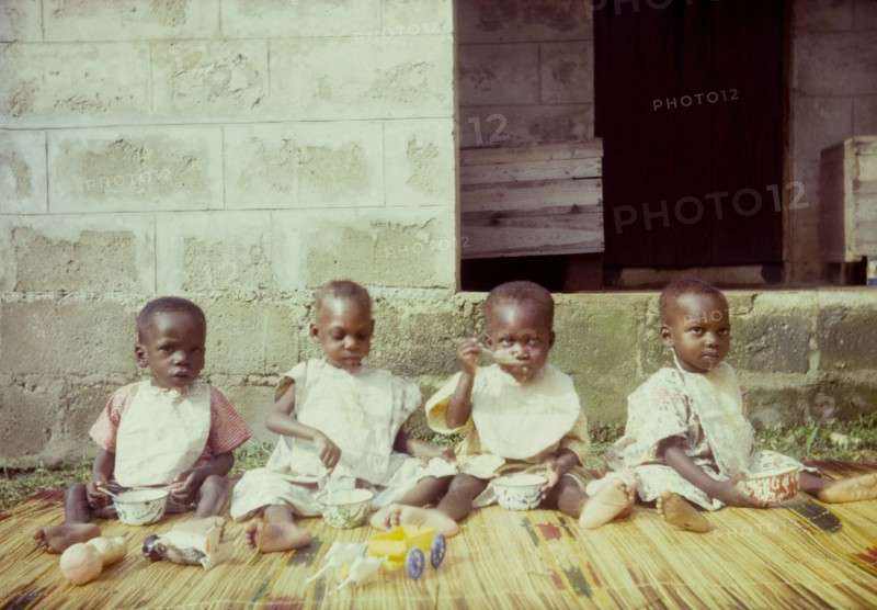 Orphans at a Church of Nigeria hospital. Four young orphans, seated on a rush mat, feed themsleves with bowls and spoons outside a Church of Nigeria hospital. Nigeria, circa 1962. Nigeria, Western Africa, Africa.