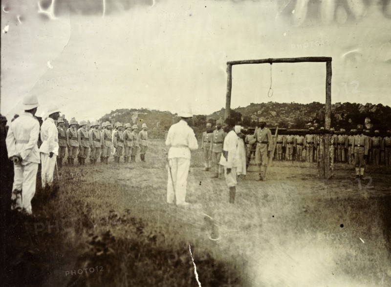 Governor reads death sentence. A European official reads a death sentence, facing a large wooden scaffold with a noose hanging from it. He is surrounded on several sides by uniformed men, both European and African. German East Africa (Tanzania), 1906. Tanzania, Eastern Africa, Africa.