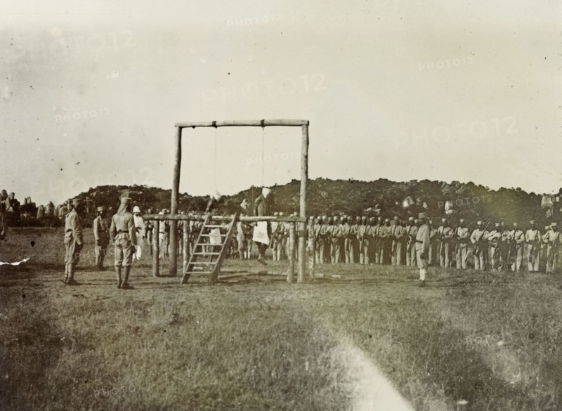 Colonial execution. The bodies of two African men hang from a wooden scaffold, their execution having been witnessed by colonial troops and officers. German East Africa (Tanzania), 1906. Tanzania, Eastern Africa, Africa.