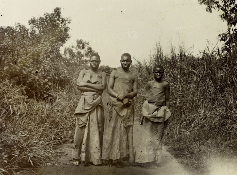 Local people, Uganda, 1906. A woman, man and girl pose for their photograph on a track by high vegetation. All are wearing long garments made of bark cloth. Uganda, 1906. Uganda, Eastern Africa, Africa.