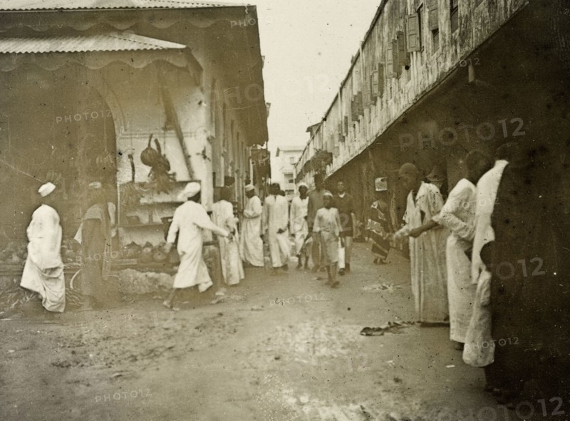 Zanzibar 'business quarter'. Men in long robes walk purposefully along a busy, narrow road in the centre of Zanzibar, originally captioned as the 'business quarter'. A shop on the left can be seen selling fruit and vegetables. Zanzibar (Tanzania), 1906. Zanzibar Town, Zanzibar Urban/West, Tanzania, Eastern Africa, Africa.