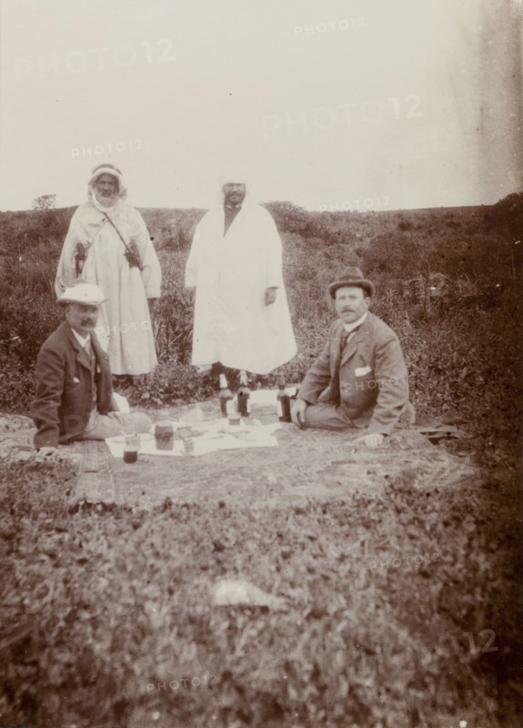 Picnic lunch, Morocco. Two European men enjoy a picnic on a blanket in an open field: their Moroccan guides standing behind them. The European man on the right is Frederick Stanbury, the other, his travelling companion in Morocco, 'Hunter'. The photograph was taken on an excursion to archaeological ruins. Morocco, 1898. Morocco, Northern Africa, Africa.