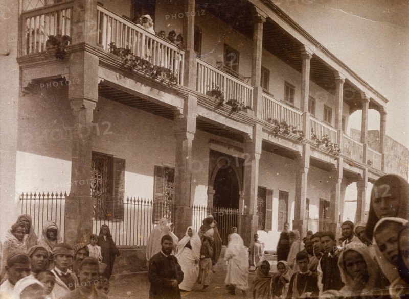 Russi's house' in Safi. Crowds of people stare in wonderment at the camera beside a large colonial-style building identified as 'Russi's House'. The building has two storeys, a long wooden balcony running across the first floor and shuttered windows. Safi, Morroco, 1898. Safi, Safi, Morocco, Northern Africa, Africa.