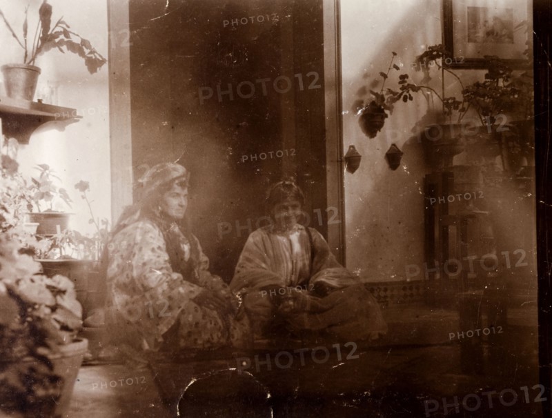 Mrs Russi dressed as a 'moorish woman'. Mrs Russi, a European woman, sits in the doorway of a colonial house dressed up as a 'moorish woman'. Clothed in a robe, she wears her hair (or wig) long and loose and is accompanied by a Moroccan girl whio sits cross-legged by her side. Safi, Morocco, 1898. Safi, Safi, Morocco, Northern Africa, Africa.