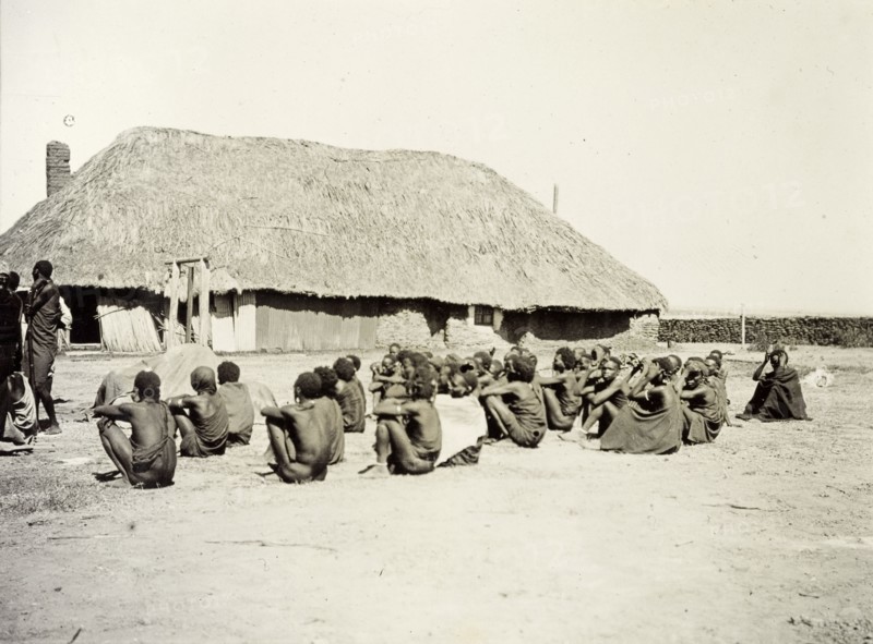 Depot for hiring porters. A group of African men and boys sit outside a large, thatched building, waiting to be engaged as porters and carriers. German East Africa (Tanzania), 1906. Tanzania, Eastern Africa, Africa.