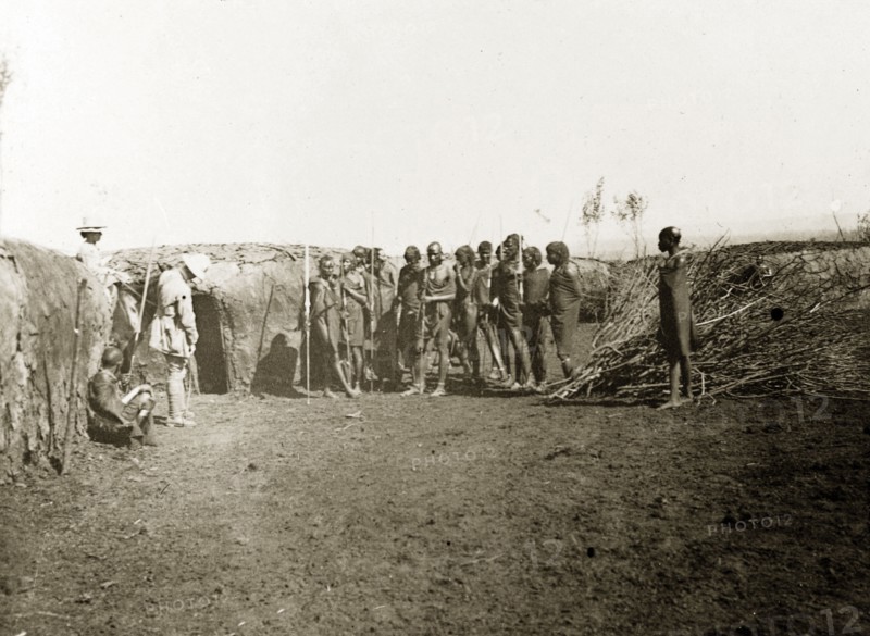 Inside a Maasai village. A group of Maasai watch as two European men examine a wall made from mud inside their village. British East Africa (Kenya), 1906. Kenya, Eastern Africa, Africa.