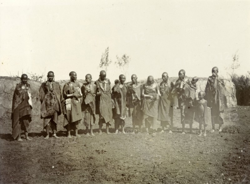 Maasai adults and children. A group of Maasai adults and children wearing traditional dress and jewellery line up for the camera. Behind them can be seen the low, breadloaf-shaped houses typical of Maasai communities. British East Africa (Kenya), 1906. Kenya, Eastern Africa, Africa.