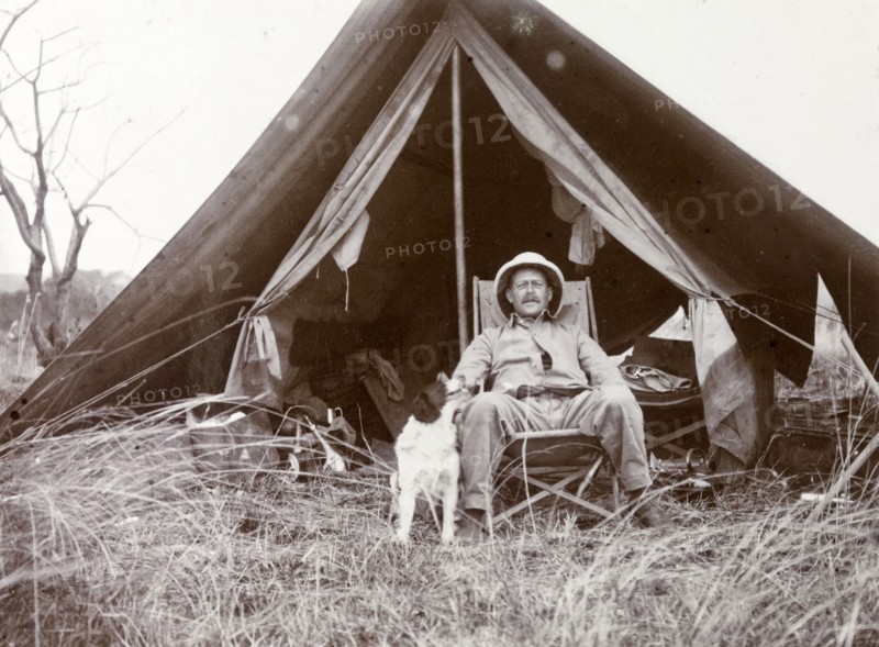 Frederick Stanbury outside a tent. Stanbury is seated on a canvas chair in front of a small open tent, a pet dog by his side. Near Lake Baringo, British East Africa (Kenya), 1906., Rift Valley, Kenya, Eastern Africa, Africa.