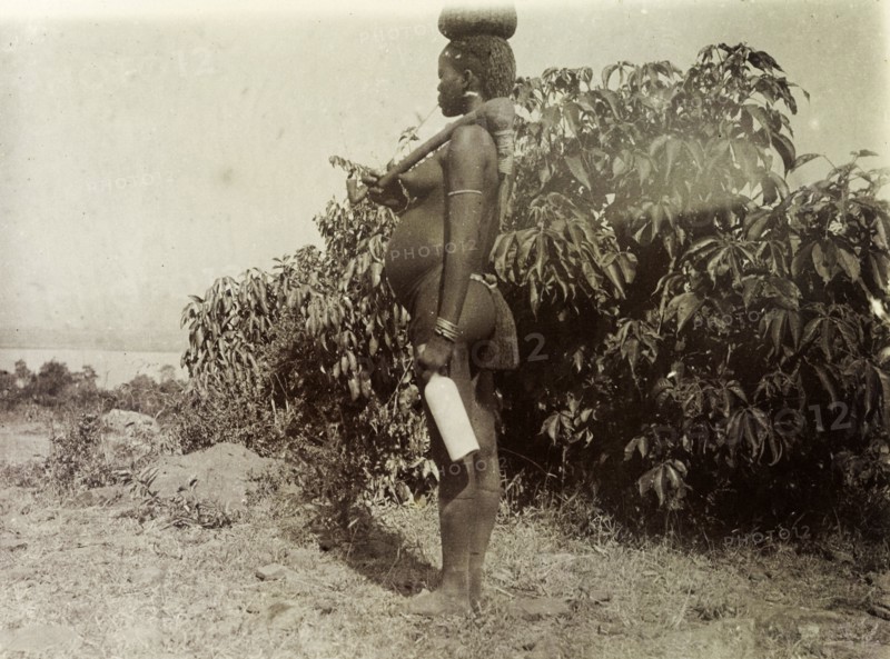 Kavirondo woman. Profile shot of a naked Kenyan woman carrying a bottle of milk on her way to a local market. She rests a hand tool over her shoulder and balances another pot on her head. Kavirondo, British East Africa (Kenya), 1906. Kavirondo, Nyanza, Kenya, Eastern Africa, Africa.