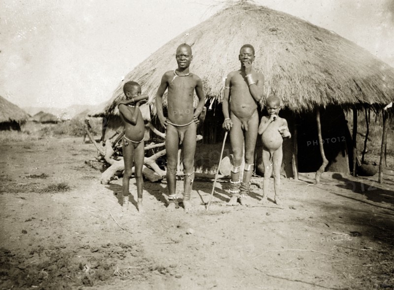 Luo men and children. Two nearly-naked men and two children pose for the camera outside a round, grass-roofed dwelling. Near Kisumu, British East Africa (Kenya), 1906., Nyanza, Kenya, Eastern Africa, Africa.