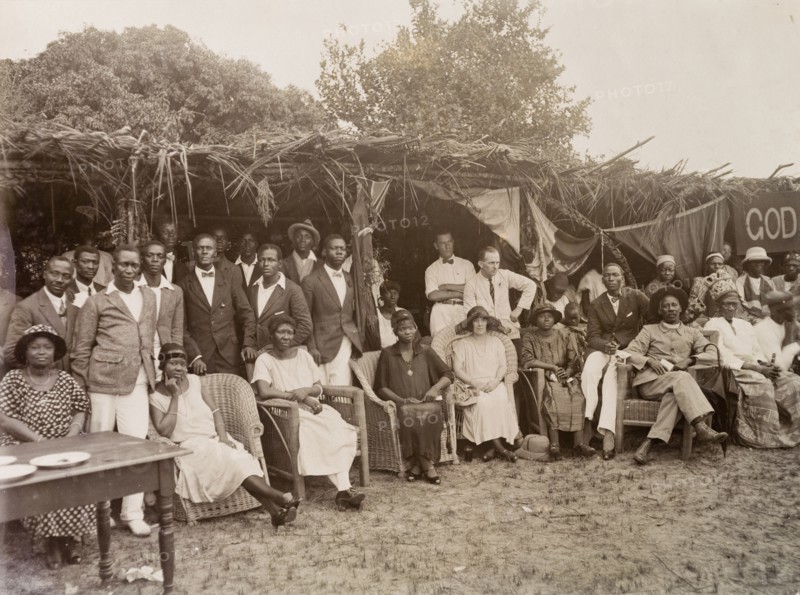 Empire Day celebrations. Informal group portrait of Africans and Europeans at Empire Day celebrations at Badragry. The group includes European employees of Miller Brothers and the African Oil Nuts Company and an African priest, probably an Anglican. A partially obscured banner reads 'God Save the King'. Nigeria, May 1924. Nigeria, Western Africa, Africa.