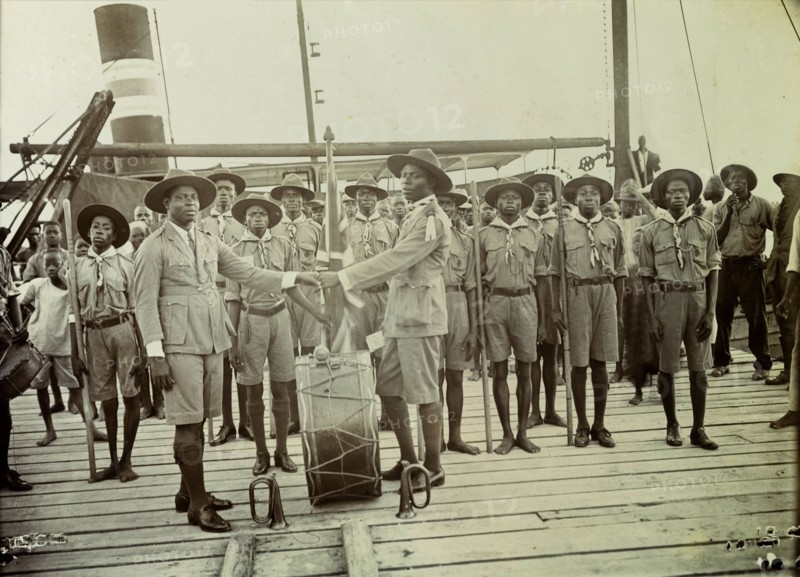 Boy scouts. Change of watch or duty between two Nigeria Boy Scout leaders onboard a steamer. The two Scout masters shake hands across a large kettle drum that stands between them, watched by an audience of at least 20 African Boy Scouts who are all standing to attention. Nigeria, 1928. Nigeria, Western Africa, Africa.