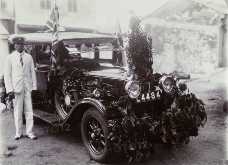 Remembrance Day car. An African chauffeur stands beside a car decorated with foliage and Union flags for Remembrance Day. Nigeria, circa 1925. Nigeria, Western Africa, Africa.
