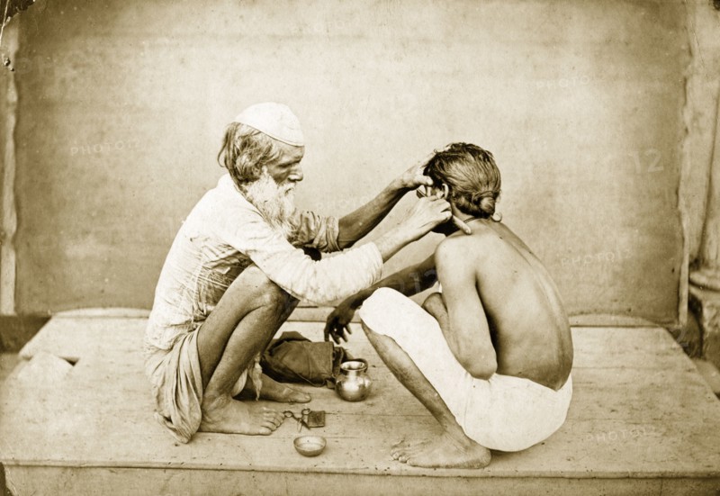 An Indian barber. An Indian barber crouches down on an outdoor platform as he shaves a customer's face. Probably Bengal, India, circa 1900. India, Southern Asia, Asia.