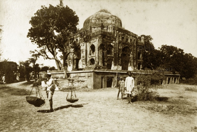 Ruined mosque, Serampore. A man carries baskets on a pole over his shoulder at the site of a ruined mosque. The stonework of the mosque is crumbling, the walls overgrown with vegetation. Serampore, India, circa 1890. Serampore, West Bengal, India, Southern Asia, Asia.