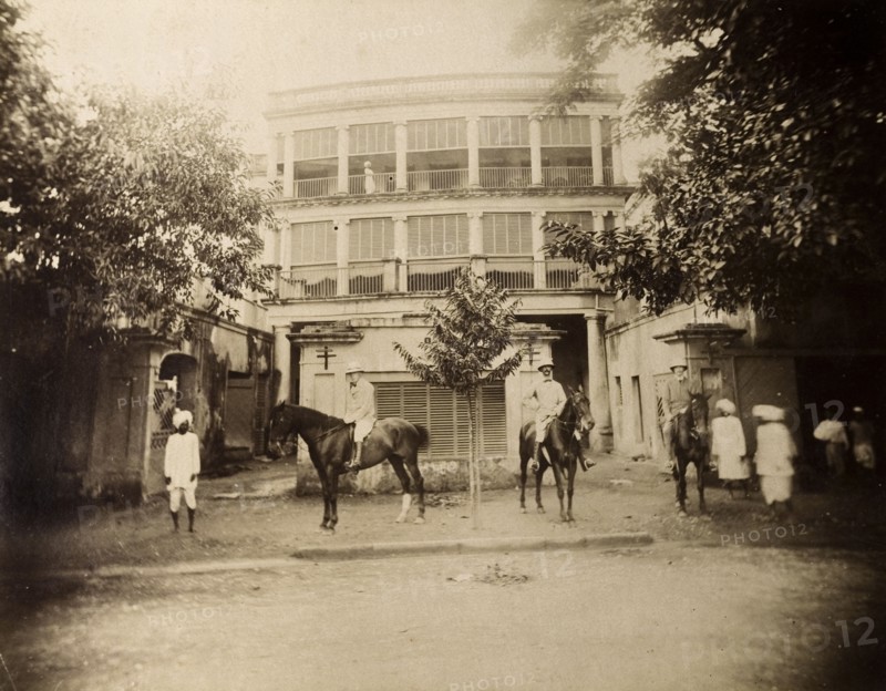 Saddling up outside a colonial house. S.A.H. Sitwell and friends pose on horseback in the walled compound of a multi-storeyed colonial house with wide verandas and blinds. Several Indian servants are also present. Calcutta (Kolkata), India, August 1895. Kolkata, West Bengal, India, Southern Asia, Asia.