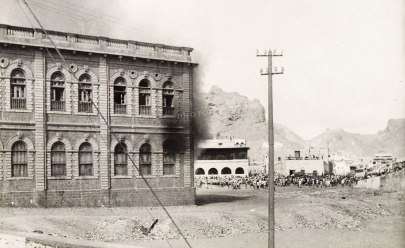 Building set on fire during the Arab riots in Aden, 1947. Black smoke billows from a building in the Jewish quarter of Aden, while crowds of people can be seen milling about at a road junction, during the Arab riots that took place between 1-3 December 1947 in response to the United Nations Partition Plan for Palestine. Aden, Yemen, December 1947. Aden, Adan, Yemen, Middle East, Asia.