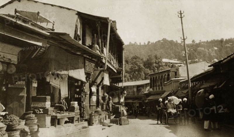 Bazaar at Mussoorie, India. Ramshackle buildings line a winding road at a bazaar in Mussoorie. A European woman visits the market stalls, pulled along in a rickshaw by uniformed Indian servants. Mussoorie, India, circa 1890. Mussoorie, Uttaranchal, India, Southern Asia, Asia.