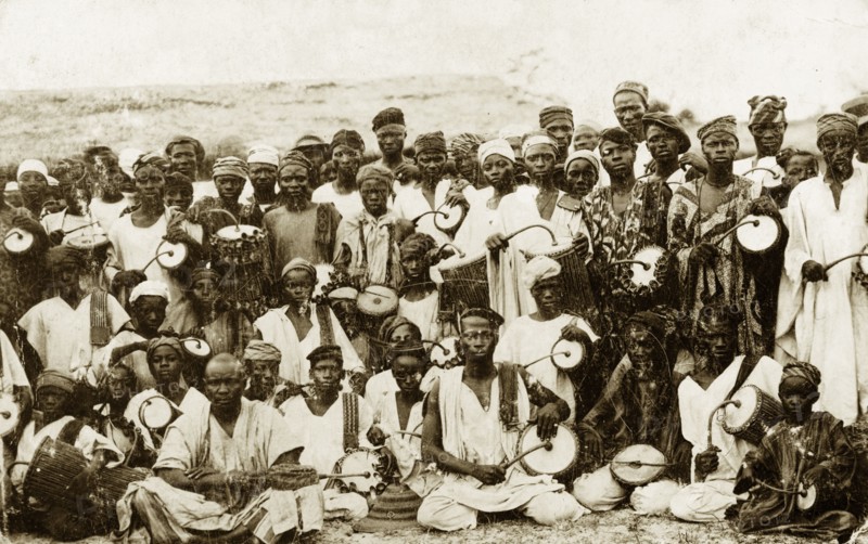 Tom-tom drummers. A group of dancers and tom-tom drummers pose for the camera holding curved drumsticks poised over their instruments. Nigeria, circa 1928. Nigeria, Western Africa, Africa.