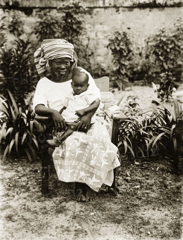 Mother and child, Nigeria. Portrait of a seated African woman holding a baby on her knee. She wears traditional clothing including a head scarf and a patterned wrap-around skirt. Nigeria, circa 1925. Nigeria, Western Africa, Africa.