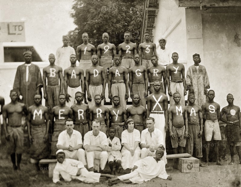 Christmas photograph of staff at the African Oil Nuts Company and Miller Brothers. Three rows of barechested African workers pose for the camera, each man's chest painted with a letter to spell out '1923, Badagry, Merry Xmas'. Four Europeans dressed in white, sit on a makeshift bench up front beside three African children, possibly domestic servants. Badagry, Nigeria, circa 1923. Badagry, Lagos, Nigeria, Western Africa, Africa.
