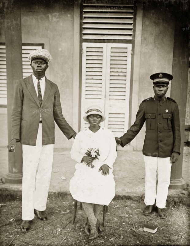 Portrait of Salvation Army workers. Two African men in Western dress stand on either side of a seated woman holding a bunch of flowers. The man on the right wears a Salvation Army cap and jacket, the woman wears a long-sleeved dress, thick tights, and high-heeled leather shoes. She also wears a Salvation Army hat. Badagry, Nigeria, circa 1928. Badagry, Lagos, Nigeria, Western Africa, Africa.