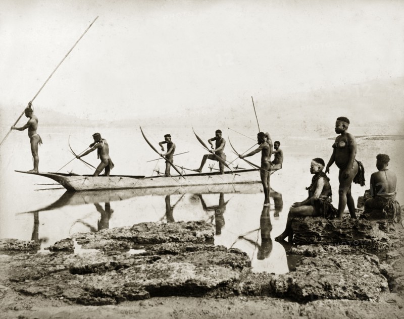 Andaman Islanders fishing. Six nearly naked men stand in a canoe, or in shallow water beside it, poised with bows and arrows ready to catch fish. Three women sit or stand on rocks at the water's edge. Andaman Islands, India, circa 1900. India, Southern Asia, Asia.