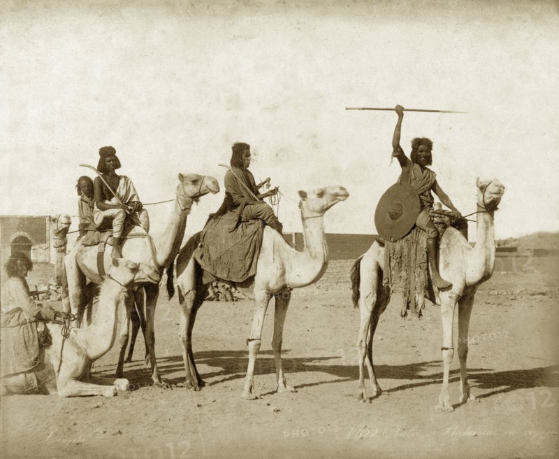 Bisharin men on camels. Five Bisharin men armed with swords, spears and shields pose seated on their camels, one of which is sitting down. Probably Sudan, North Africa, circa 1885. Sudan, Eastern Africa, Africa.