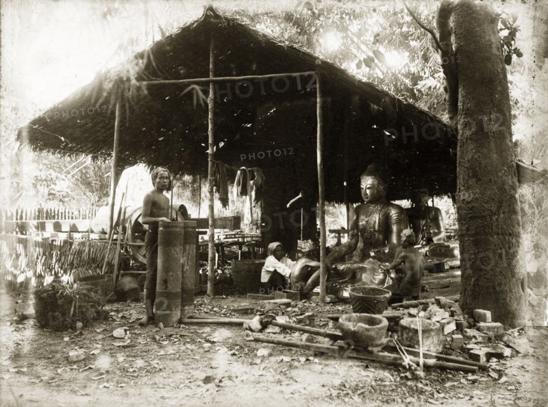 Idol-making, Burma (Myanmar). Two craftsmen sit, under a thatched, open-walled shelter, burnishing or painting a large cross-legged statue of the Buddha. A woman squats beside them. A second statue is visible in the background: a collection of metalworking tools in foreground. Burma (Myanmar), circa 1900. Burma (Myanmar), South East Asia, Asia.
