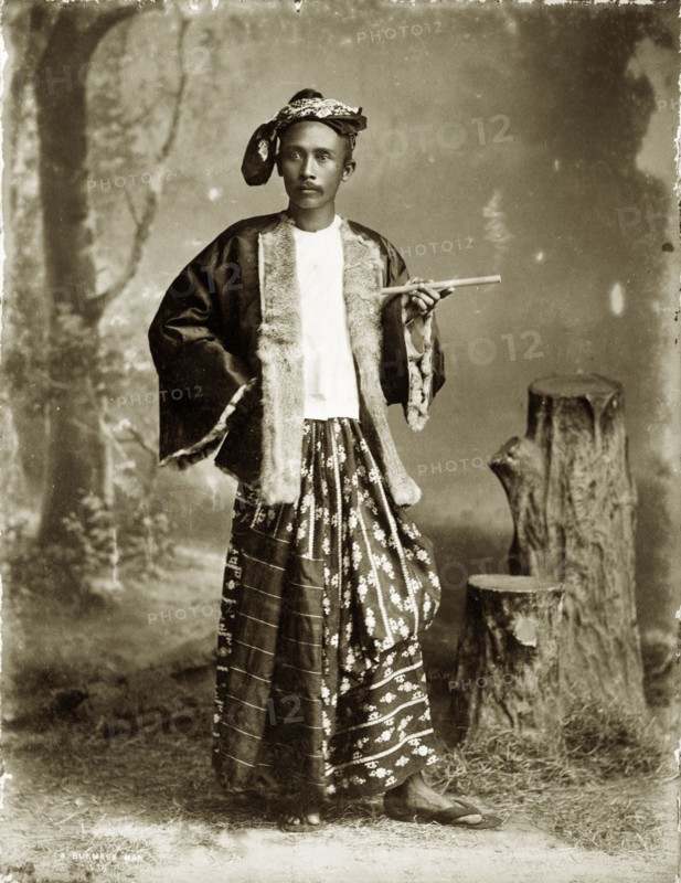 Portrait of a Burmese gentleman. A posed studio portrait of a finely-dressed Burmese gentleman. His outfit comprises a fur-lined silk jacket, headscarf and a traditional silk 'longyi' or wraparound skirt. He poses for the camera holding a smoking cheroot (a type of cigar). Burma (Myanmar), circa 1885. Burma (Myanmar), South East Asia, Asia.