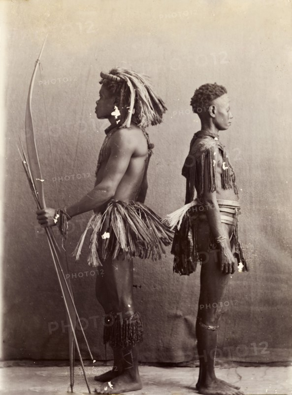 Andaman Islanders. A man and a woman from the Andaman Islands stand, back to back in profile, against an unpainted backdrop. The man holds a bow and long arrows: both wear grass-like aprons and neckpieces. Andaman Islands, India, circa 1900., Andaman and Nicobar Islands, India, Southern Asia, Asia.