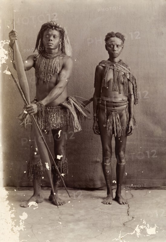 Couple from the Andaman Islands. A man and a woman from the Andaman Islands stand, facing the camera, against an unpainted backdrop. The man holds a bow and long arrows: both wear grass-like aprons and neckpieces. Andaman Islands, India, circa 1900., Andaman and Nicobar Islands, India, Southern Asia, Asia.