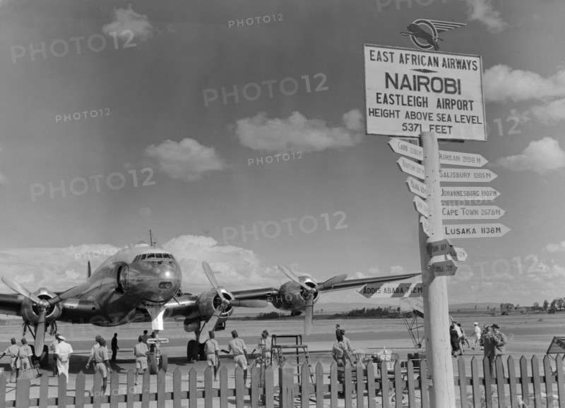 Eastleigh Airport, Nairobi. Airport staff mill around a double-propellered passenger plane that sits on the runway at Eastleigh airport. A sign post on the right indicates the distance in miles to various African cities. Nairobi, Kenya, 9 November 1952. Nairobi, Nairobi Area, Kenya, Eastern Africa, Africa.