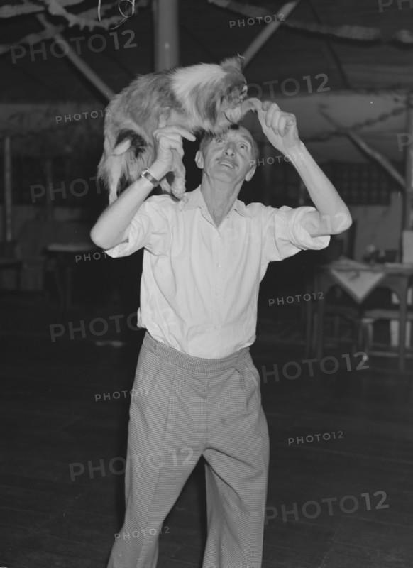Tom dances with a dog. A man identified as 'Tom', lifts a worried-looking collie dog onto his shoulder at a Christmas party being held at the Forest Inn bar. Possibly Nairobi, Kenya, 28 December 1952. Kenya, Eastern Africa, Africa.