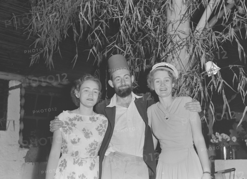Charles Trotter at a Christmas party. Charles Trotter, a professional photographer whose work is widely represented in this collection, enjoys himself at a Christmas party. He sports a fake fez hat and poses with two women identified as 'Barbara and Mary' outside the Forest Inn bar. Possibly Nairobi, Kenya, 25 December 1952. Kenya, Eastern Africa, Africa.