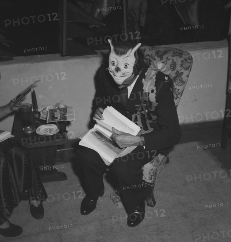 Ron Richardson with mask. Ron Richardson takes a break from the merriment at the Archer's New Year's party. Decorated with a paper garland, he sits engrossed in a set of papers, his fancy dress mask pushed up on top of his head. Kenya, 31 December 1952. Kenya, Eastern Africa, Africa.