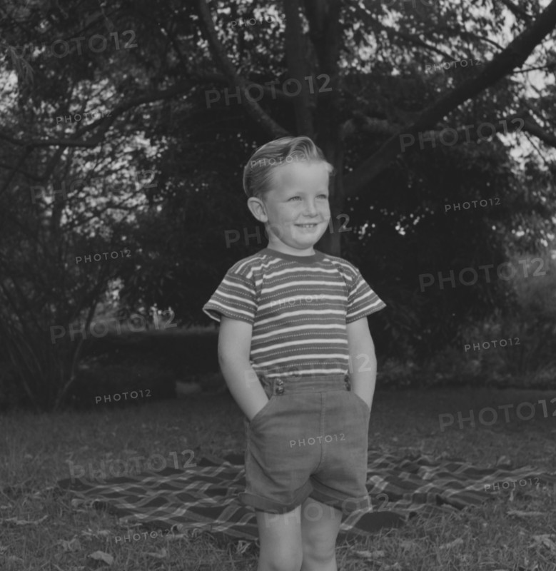 Cheerful boy, Kenya. Stephen Ward stands, hands in pockets, smiling broadly beside a picnic rug laid out in a garden. Kenya, 1 January 1953. Kenya, Eastern Africa, Africa.