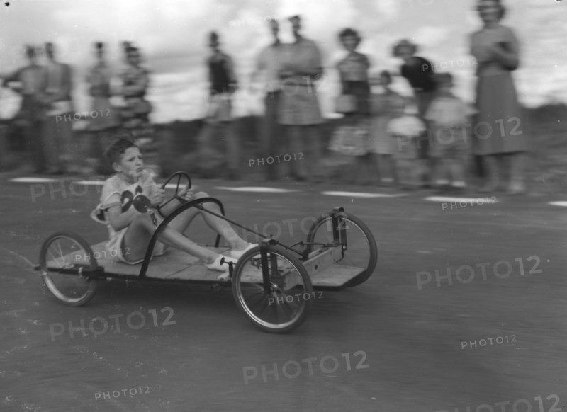 Number 26 in the soap box derby. A young boy braces himself as he whizzes past crowds riding a homemade go-cart, entry number 26 in a soap box derby race. Kenya, East Africa, 03 January 1953. Kenya, Eastern Africa, Africa.
