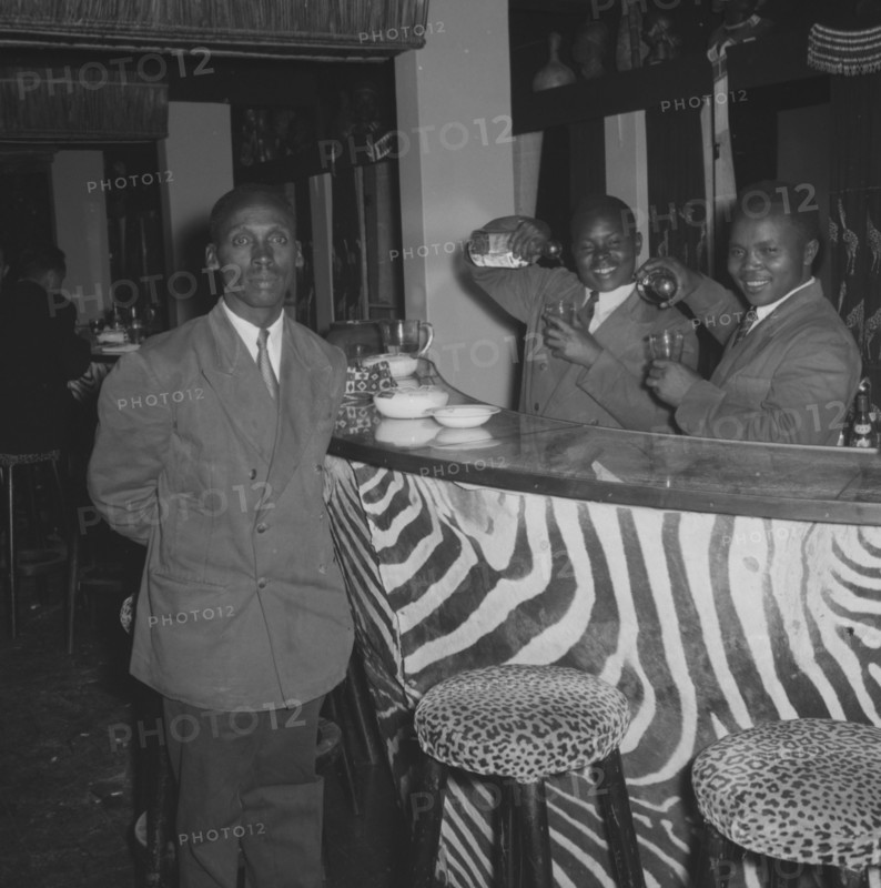 Bartenders at the Equator Club. Portrait of three uniformed waiters at the Equator Club. Two African bartenders jokingly pour drinks into glasses behind a bar decorated with zebra stripes and leopard-print stools. Nairobi, Kenya, 21 September 1957. Nairobi, Nairobi Area, Kenya, Eastern Africa, Africa.