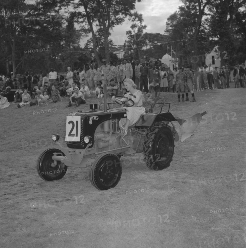 Tractor in a vehicle parade. A young woman with coiffeured hair and heels, drives a small tractor past a crowd of onlookers in a vehicle parade at the Royal Show. Kenya, 26 September 1957. Kenya, Eastern Africa, Africa.