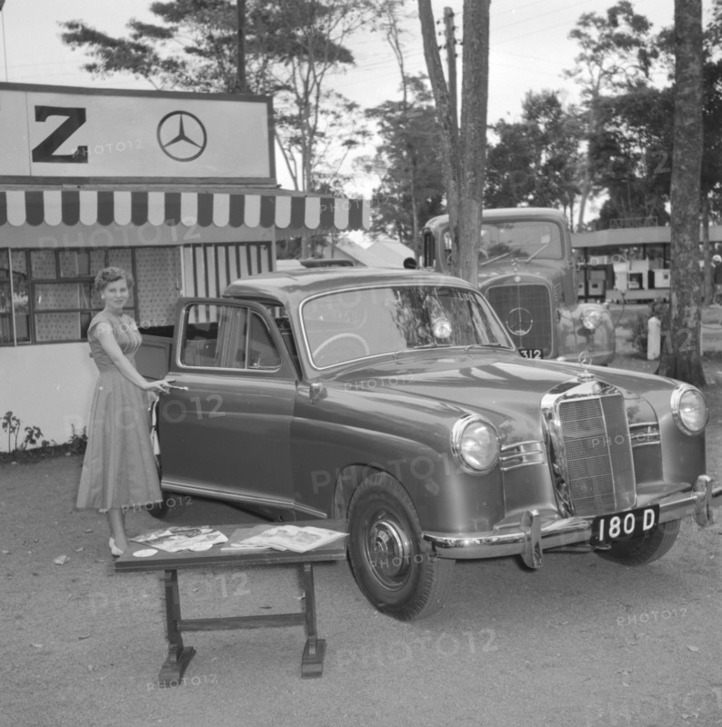 D.T. Dobie stand. A woman poses for the camera, opening the door of a new Mercedes car on display at the D.T. Dobie stand at the Royal Show. Kenya, 25 September 1957. Kenya, Eastern Africa, Africa.
