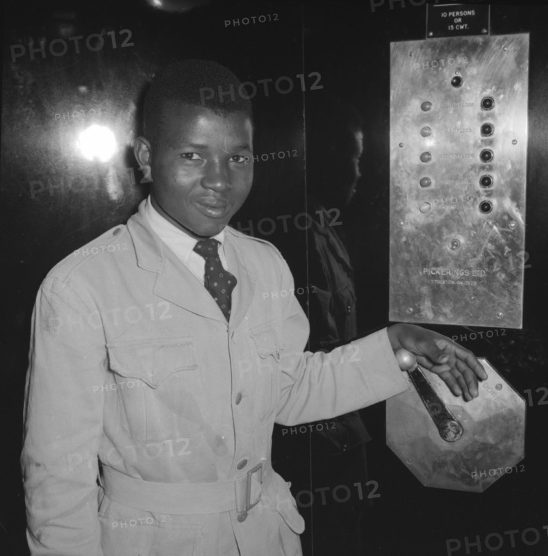 Congress House lift attendant. A young African lift attendant smiles for the camera, his hand resting on a manual handle inside an elevator at Congress House. Nairobi, Kenya, 11 October 1957. Kenya, Eastern Africa, Africa.