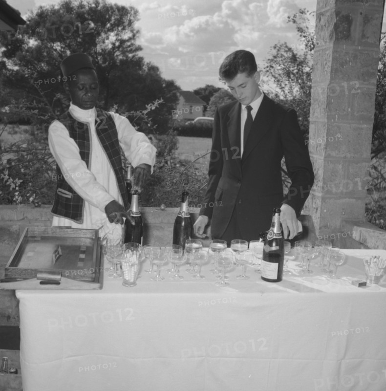 Wedding barmen. A young European man assists a uniformed African servant to pour champagne at the Hodgson-Wilkinson wedding. Kenya, 12 October 1957. Kenya, Eastern Africa, Africa.